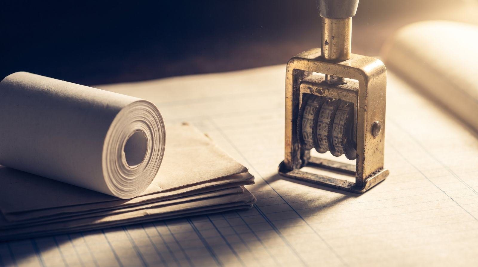 A brass date stamp standing on ruled ledger paper beside a roll of receipt paper
